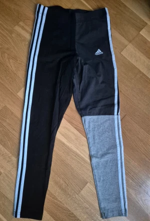 Adidas leggings med ränder - Snygga Adidas leggings med klassiska vita ränder längs sidorna. Ena benet är svart och det andra är delvis grått, vilket ger en cool kontrast. Adidas-loggan finns på höften. Perfekta för träning eller chill. Stretchigt material för skön passform.