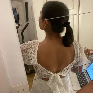 Vit spetsklänning med öppen rygg - Superfin vit kortklänning i spets med puffiga kortärmar och omlott-v-ringning. Klänningen har volang nedtill och en snygg öppen rygg med knytband. Perfekt som avslutningsklänning och student! Använd endast en gång!