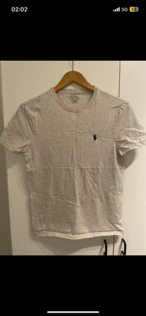 Ljusgrå t-shirt från Polo Ralph Lauren - Snygg ljusgrå t-shirt från Polo Ralph Lauren i bomull med klassisk rund hals och korta ärmar. Custom slim fit-modell med den ikoniska blå Polo-loggan broderad på bröstet. Perfekt basic att ha i garderoben.