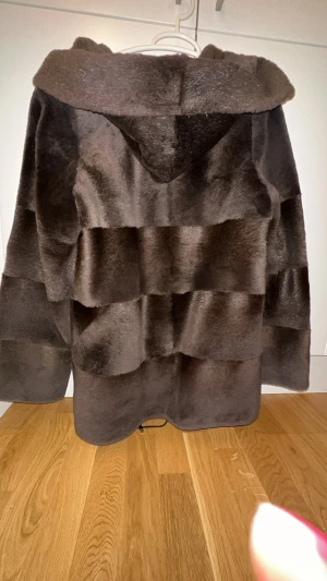 Äkta Bäverpäls / pälsjacka - M/S - Säljer en brun äkta pälsjacka från Samar med stor huva och breda horisontella paneler. Jackan har knappar framtill och ett rött innerfoder. Den är långärmad och har en loose passform, perfekt för dig som vill ha en varm och snygg look under vintern.