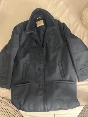 Mörkblå rock från Billrock - Säljer en stilren mörkblå rock från Belrock med klassisk krage och quiltat foder. Jackan har fyra fickor framtill, knäppning med knappar och detaljerad krage med stickad insida. Materialet är polyester och jackan har en rak, normal passform. Obs kan ha små defekter