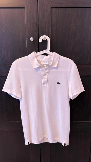 Vit pikétröja från Lacoste - Snygg vit pikétröja från Lacoste med klassisk krage och korta ärmar. Passform slim fit, Tröjan har två knappar framtill och den ikoniska gröna krokodilloggan på bröstet. Perfekt för dig som gillar stilrena och tidlösa plagg.