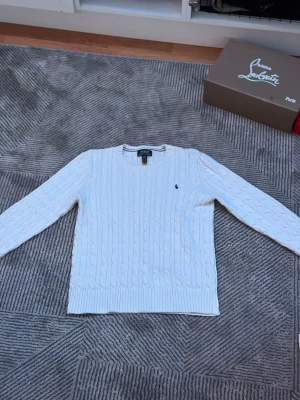 Vit kabelstickad tröja Polo Ralph Lauren - Vit kabelstickad tröja från Polo Ralph Lauren med klassisk rund hals och broderad logga på bröstet. Tröjan har långa ärmar och ribbade muddar vid ärmslut och nederkant. Perfekt för dig som gillar stilrena och tidlösa plagg. PS: ANVÄND 1 GÅNG OCH TVÄTTAD 1 GÅNG