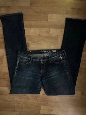 Replay jeans bootcut blå W28 L34 - Snygga mörkblå jeans från Replay med klassisk bootcut och låg midja. Jeansen har fem fickor, detaljerade sömmar och ett läderpatch med Replay-logga bak. Passar dig som gillar en tidlös look med lite extra attityd.