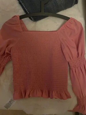 Rosa blus med volang från Vero Moda - Supersöt rosa blus från Vero Moda i storlek S. Smockad kropp med volangkant nedtill och puffiga långärmar med smock vid ärmslut. Fyrkantig halsringning och mjukt, lätt material som känns skönt mot huden. Perfekt för dig som gillar romantiska detaljer och trendig stil.