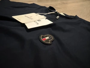Mörkblå t-shirt från Moncler - Snygg mörkblå t-shirt från Moncler med klassisk rund hals och korta ärmar. På bröstet sitter ett broderat Moncler-emblem i svart, vitt och rött. Materialet är mjuk bomull som känns skönt mot huden. Perfekt för dig som gillar stilrena och exklusiva plagg.