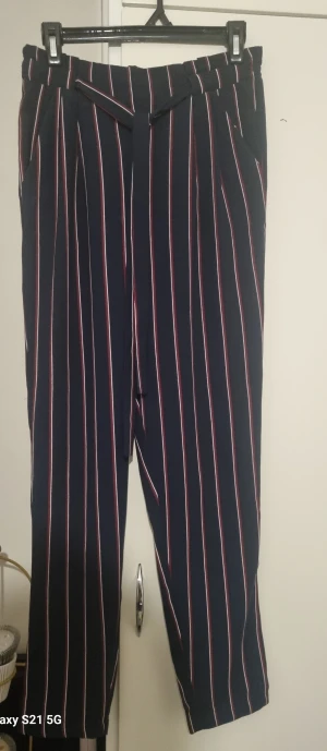 Mörkblå randiga byxor med bälte - Snygga mörkblå byxor med vertikala vita och röda ränder. Byxorna har ett medföljande bälte i samma tyg och en rak, lite loose passform. Perfekt för dig som gillar en trendig och bekväm stil. Midjan är normal och materialet känns mjukt och följsamt.