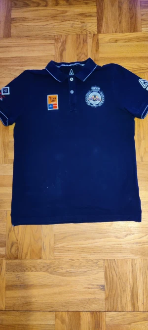 Mörkblå pikéskjorta Gaastra XL - Snygg mörkblå pikéskjorta från Gaastra i bomull, med broderade och tryckta detaljer från Copa del Rey. Skjortan har korta ärmar, vit kontrast på krage och ärmslut, samt flera coola patchar och knappar framtill. Perfekt för dig som gillar nautisk stil och sportiga vibbar.