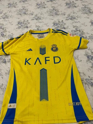 Al Nassr Ronaldo 7 matchtröja Adidas - Gul och blå Al Nassr matchtröja från Adidas med Ronaldo 7 på ryggen. Tröjan har korta ärmar, klubbmärke och sponsortryck framtill samt klassiska Adidas-ränder på axlarna. Tillverkad i lätt polyester, perfekt för fotboll och sport.