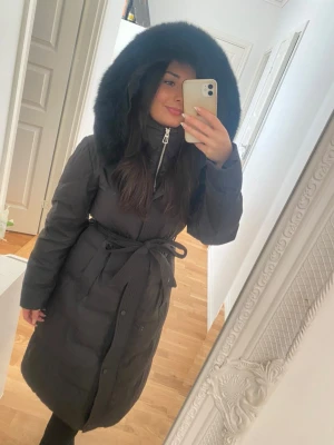 Svart lång vinterjacka med päls - Säljer en svart lång dunjacka med fluffig päls på huvan och snyggt bälte i midjan. Jackan har dragkedja och knappar framtill, quiltad design och långa ärmar. Perfekt för kalla dagar. Storlek 34 men lite stor i storlek så passar utan tvekan en 36 med. Endast använd några ggr. Mitt pris 2700kr , ord pris 3999kr 