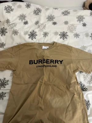 Beige Burberry t-shirt med tryck - Snygg beige t-shirt från Burberry med klassiskt svart logotryck 'BURBERRY LONDON ENGLAND' på bröstet. T-shirten har rund hals och korta ärmar, perfekt för en clean och stilren look. Materialet är mjuk bomull som känns skönt mot huden.