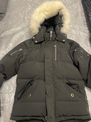 Svart vinterjacka med päls från Moose Knuckles - Svart dunjacka från Moose Knuckles med fluffig vit päls på huvan, coola metalldetaljer och logga på ärmen. Jackan har flera fickor med dragkedja och knappar, samt en hög krage som skyddar mot kyla. Perfekt för dig som vill ha en stilren och varm vinterjacka.. passar som S/M