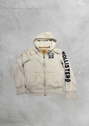 Vintage Hollister zip hoodie🏄‍♂️ - Vit hoodie från Hollister med dragkedja och huva. Stor tryckt text 'HOLLISTER' i mörkblått längs ena ärmen och 'SO CAL 22' på bröstet. Mjuk bomullskänsla, fickor fram och snörning i huvan. Perfekt för chill dagar och streetstyle. Storlek L men sitter som s/m