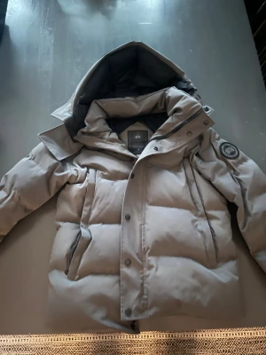 Grå dunjacka Canada Goose Fusion S - Säljer en grå Canada Goose dunjacka Fusion i storlek S. Jackan har huva med avtagbar päls, dragkedja och knappar framtill samt två sidofickor. Klassisk Canada Goose-märke på ärmen och snygg puffad design som håller dig varm hela vintern.