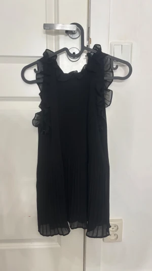 Svart volangklänning från Zara - Supersöt svart blus från Zara med volangdetaljer längs axlarna och plisserad nederdel. Ärmlös modell i lätt och luftig polyester som ger en snygg siluett