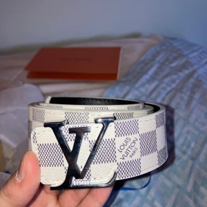 L.V rutig skinnbälte - Säljer ett L.V bälte i skinn med klassiskt rutigt mönster i vitt och grått. Bältet har en stor, silverfärgad L.V-logga som spänne och kommer i originalförpackning och hålgörare. Perfekt accessoar för att lyfta din outfit med lyxig vibe. Kvitto och såvidare kommer med hör av dig vid första fundering
