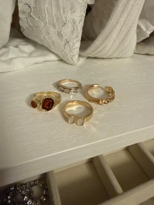 Fyra chunky ringar i guld  - Säljer ett set med fyra chunky ringar i guld. En ring har två vita stenar, en har en stor röd sten och en mindre, en är guldfärgad med tre vita pärlor och en har en twistad design med små stenar. Perfekt för dig som gillar statement-smycken.