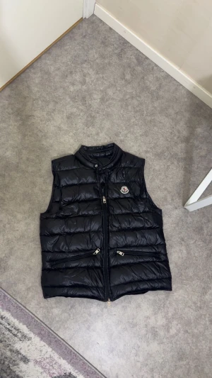 Svart Moncler pufferväst med dragkedja - Svart pufferväst från Moncler med glansig finish och klassisk logga på bröstet. Västen har två snedställda fickor med dragkedja och hög krage med knapp. Perfekt att slänga över en hoodie för extra värme under höst och vinter. OBS! Den har ett litet hål bara