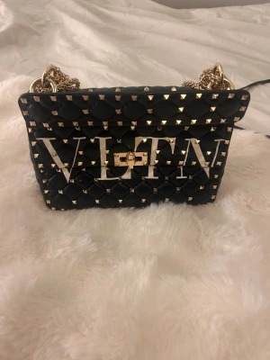 Valentino svart axelväska med nitar - Snygg svart axelväska från Valentino med stora VLTN-bokstäver framtill. Väskan är quiltad i skinn och dekorerad med guldiga nitar och kedja. Låsdetalj i guld och cool design som sticker ut. Perfekt för dig som vill ha en statementväska.