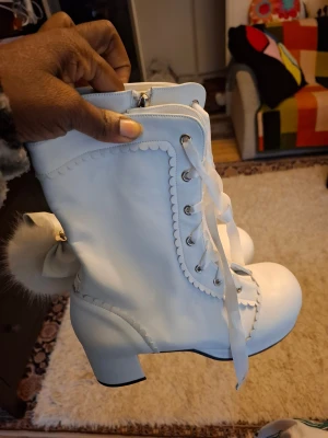 Vita boots med rosett och pälsboll - Supersöta vita boots med chunky klack, snörning med breda band och dekorativ scallop-kant. På sidan sitter en fluffig pälsboll och rosett som ger extra kawaii-vibe. Gjorda i syntetmaterial med dragkedja på insidan för enkel påtagning.