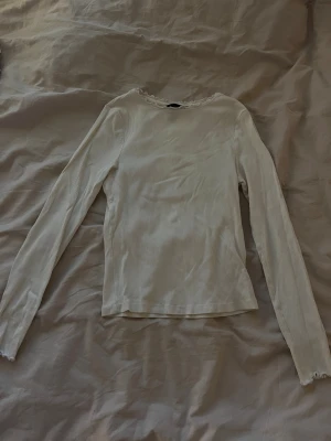 Vit ribbad långärmad topp - Säljer en vit långärmad topp från New Look med ribbad struktur och söt vågig kant vid halsringning och ärmslut. Modellen är slim och sitter snyggt mot kroppen. Perfekt att styla med jeans eller kjol för en clean look. Ett litet igensytt hål på höger arm.