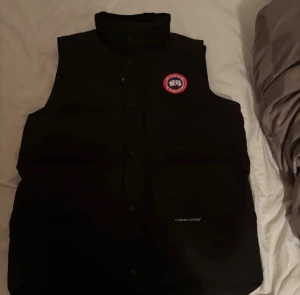 Svart dunväst från Canada Goose - Säljer en svart dunväst från Canada Goose med klassisk logga på bröstet. Västen har hög krage, tryckknappar framtill och diskret Canada Goose-brodyr på ena sidan. Perfekt för lager-på-lager och riktigt clean look.