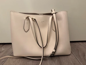 Beige axelväska från Kate Spade - Snygg beige axelväska från Kate Spade med stilren design och praktiska handtag. Väskan har ett stort huvudfack och en innerficka med dragkedja. Tillverkad i slätt skinn med ljusa detaljer och diskret logga framtill. 