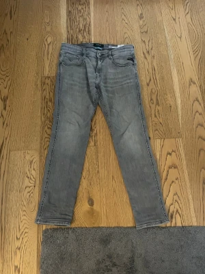 Replay Anbass grå slim jeans - Snygga grå jeans från Replay, modell Anbass med slim fit. Jeansen har klassisk femficksdesign, svarta detaljer och en cool tvättad look. Materialet är jeans med stretch för extra komfort. Passar dig som gillar smalare siluett och stilren vibe.
