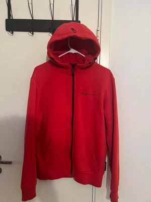 Röd hoodie från Sail Racing - Riktigt snygg röd hoodie från Sail Racing i storlek M. Tröjan har huva, dragkedja framtill och två stora fickor. Materialet är mjuk bomull och polyester, perfekt för chill dagar. Diskret logga på bröstet och cool detalj på huvan.