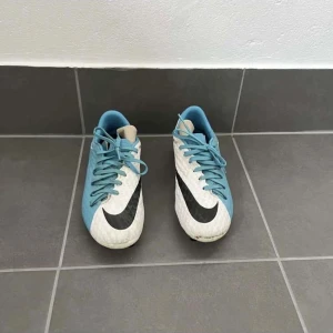 Nike fotbollsskor blå och vita - Säljer ett par Nike fotbollsskor i fräscht blått och vitt med svart swoosh på sidan. Skorna har snörning och en rund tå, tillverkade i syntetmaterial för bra grepp på planen. Perfekt för dig som vill ha snygga och funktionella skor till träningen.