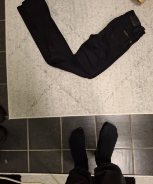 Svarta jeans från Nudie Jeans, W24 L32 - Säljer ett par svarta jeans från Nudie Jeans i modellen W24 L32. Klassisk femficksmodell med raka ben och snyggt svart tyg. Passar perfekt till dig som gillar stilrena och tidlösa jeans. Materialet är slitstarkt och bekvämt.