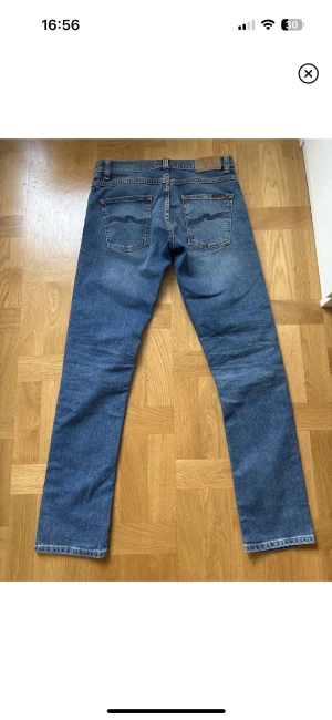 Blå jeans från Nudie Jeans, straight fit - Säljer ett par klassiska blå jeans från Nudie Jeans med straight fit och normal passform. Jeansen har fem fickor, snygga kontrastsömmar och Nudie-logga på bakfickan. Materialet är slitstark denim i bomull och färgen är mellanblå med lätt tvättad look.