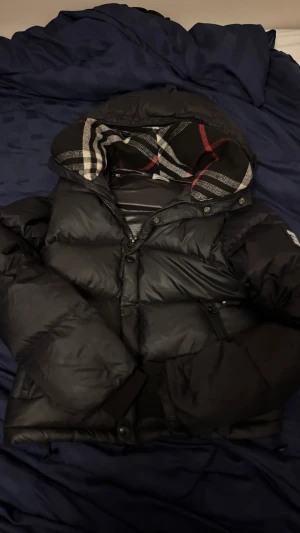 Burberry jacka / väst - Svart pufferjacka från Burberry med avtagbara ärmar så den kan användas som väst. Jackan har huva med klassiskt rutigt Burberry-mönster i svart, vitt och rött. Flera fickor med dragkedja och knappar framtill. Perfekt för kalla dagar.