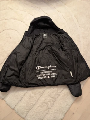 Svart pufferjacka från Champion - Säljer en svart pufferjacka från Champion med huva och snyggt broderad logga på bröstet. Jackan har dragkedja framtill, två sidofickor och quiltad design. Perfekt för dig som vill ha en stilren och varm jacka med sportig vibe.