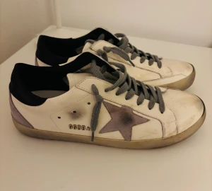Golden Goose Superstars  - Golden Goose Superstar sneakers i vitt skinn med grå snörning och en grå stjärna på sidan. Svart hälparti och slitna detaljer för en cool vintage-look. Platt sula och rund tå. Snyggt tryck på innersulan och GGDB-logga på sidan. Storlek 42, passar också 41. Har använt de ett litet tag. Komforta och passar till alla kläder. Priset kan diskuteras! Skriv i dm om ni har några funderingar. 