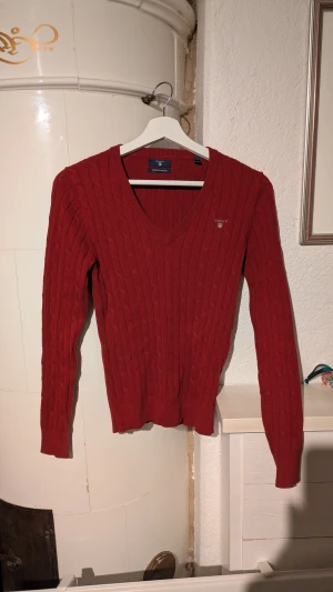 Röd stickad v-ringad tröja från GANT - Snygg röd stickad tröja från GANT med v-ringad hals och diskret logga på bröstet. Tröjan har ett klassiskt kabelstickat mönster och långa ärmar. Perfekt för dig som gillar stilrena och tidlösa plagg med lite preppy känsla.