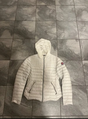 Vit dunjacka från Colmar Originals - Snygg vit dunjacka från Colmar Originals med huva och dragkedja framtill. Jackan har quiltad design, två fickor med dragkedja och en rund Colmar-logga på ärmen. Perfekt för kyliga dagar och riktigt lätt att bära.