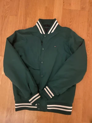 Grön bomberjacka från Tommy Hilfiger - Snygg grön bomberjacka från Tommy Hilfiger med klassiska vita ränder på krage, ärmslut och midja. Jackan har tryckknappar framtill och diskret logga på bröstet. Perfekt för dig som vill ha en stilren och sportig look.
