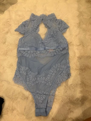 Blå spetsbody från Hunkemöller M - Supersöt ljusblå body från Hunkemöller i storlek M. Den är gjord i mesh och spets med blommiga detaljer, har korta ärmar och v-ringning. Knäppning i grenen för enkel användning.