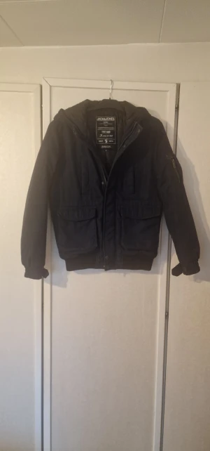Marinblå jacka från Jack & Jones - Marinblå jacka från Jack & Jones med huva, stora fickor framtill och ribbade muddar. Jackan har dragkedja och knappstängning, samt en ficka med dragkedja på ärmen. Materialet är polyester och jackan har en klassisk, loose passform. Kardborre vid ärmslut har delvis lossnat på båda sidorna, den sitter fortfarande kvar men behöver sys fast. Materialet på ovankanten av dragkedjan är slitet, färgen är något blekt på lite mer utsatta områden som kanten på fickorna och mudden är lite nopprig.