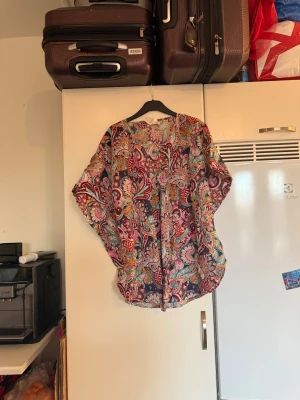 Odd Molly färgglad blus med v-ringning - Säljer en blus från Odd Molly med ett färgstarkt paisley-inspirerat mönster i rosa, blått, orange och lila. Blusen har v-ringning, lös passform och är tillverkad i 100% bomull.  Perfekt för dig som gillar boho-vibes och unika prints. Storlek one size som kanske passar mest en XL. Mycket fint skick, jag hittar inga synliga anmärkningar eller defekter. 