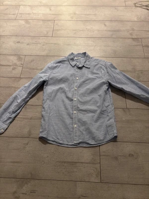 Blårandig skjorta med knappar - Snygg blårandig skjorta med klassisk krage och vita knappar framtill. Skjortan har lång ärm och är tillverkad i ett lätt bomullstyg som känns skönt mot huden. Perfekt för dig som gillar stilrena och enkla plagg med lite edge. Använd en gång!