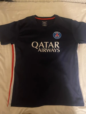 PSG fotbollströja marinblå - Snygg marinblå PSG fotbollströja med klubbmärke på bröstet och Qatar Airways tryck på framsidan. Rund hals med röd detalj på insidan och vita samt röda ränder längs sidorna. Perfekt för dig som vill representera Paris Saint-Germain.