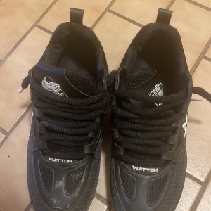 Svarta Louis Vuitton sneakers - Säljer ett par svarta Louis Vuitton sneakers med feta svarta snören, vita detaljer och tydlig logga på sidan och sulan. Skorna har coola LV-blommor på sidan och 1854-tryck bak. Materialmix av skinn och syntet, platt sula och rund tå.