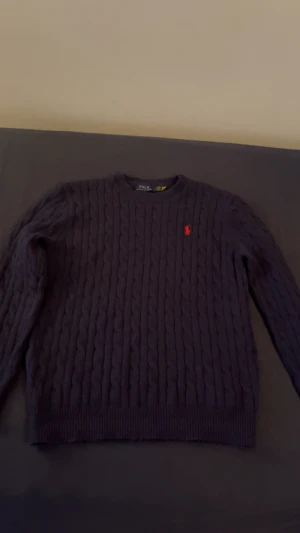 Mörkblå stickad tröja Polo Ralph Lauren - Snygg mörkblå stickad tröja från Polo Ralph Lauren i storlek L. Tröjan har ett klassiskt kabelstickat mönster och en röd broderad logga på bröstet. Rund hals och långa ärmar, perfekt för dig som gillar stilrena och tidlösa plagg.