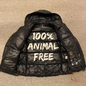Svart pufferjacka Save The Duck  - Svart pufferjacka från Save The Duck i storlek L men skulle kunna passa M. Jackan är quiltad, har huva, dragkedja framtill och två sidofickor med dragkedja. Tillverkad i syntetmaterial och är helt animal free. Orange detaljer på dragsko och patch på ärmen. Liten skada på nedre magen med ett litet hål men de är lagat så inget som märks eller man tänker på.