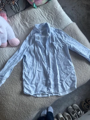 Ljusblå skjorta från A Days March - Ljusblå skjorta från A Days March i mjukt linnematerial. Klassisk design med lång ärm, bröstficka och knappar framtill. Skjortan har en avslappnad passform och är perfekt för en chill och stilren look. Passar dig som gillar enkla och fräscha plagg.