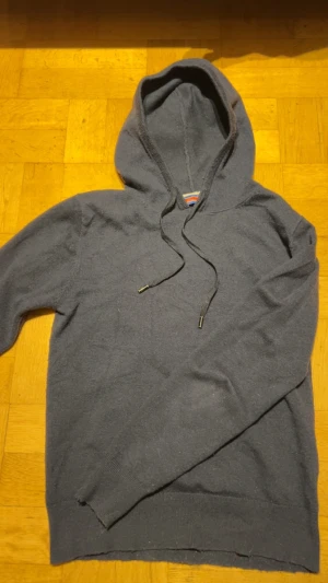 Mörkblå william de faye cashmir hoodie - Snygg mörkgrå hoodie med huva och snörning. Tröjan har långa ärmar och ribbade muddar vid ärmslut och nederkant. Perfekt för chill dagar eller när du vill ha något bekvämt och enkelt. Materialet känns mjukt och skönt mot huden.