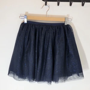 Mörkblå glittrig tyllkjol från H&M - Säljer en mörkblå kortkjol från H&M med glittrande tyll och resår i midjan. Kjolen har flera lager som ger en fluffig och voluminös look. Perfekt för dig som gillar att sticka ut med lite extra glitter i outfiten.