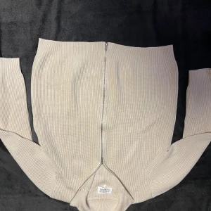 Beige stickad kofta från Maison Margiela - Säljer en beige stickad kofta från Maison Margiela med hög krage och tvåvägsdragkedja framtill. Tröjan har ribbad struktur och långa ärmar, perfekt för lager-på-lager. Klassisk och clean design som funkar till allt.